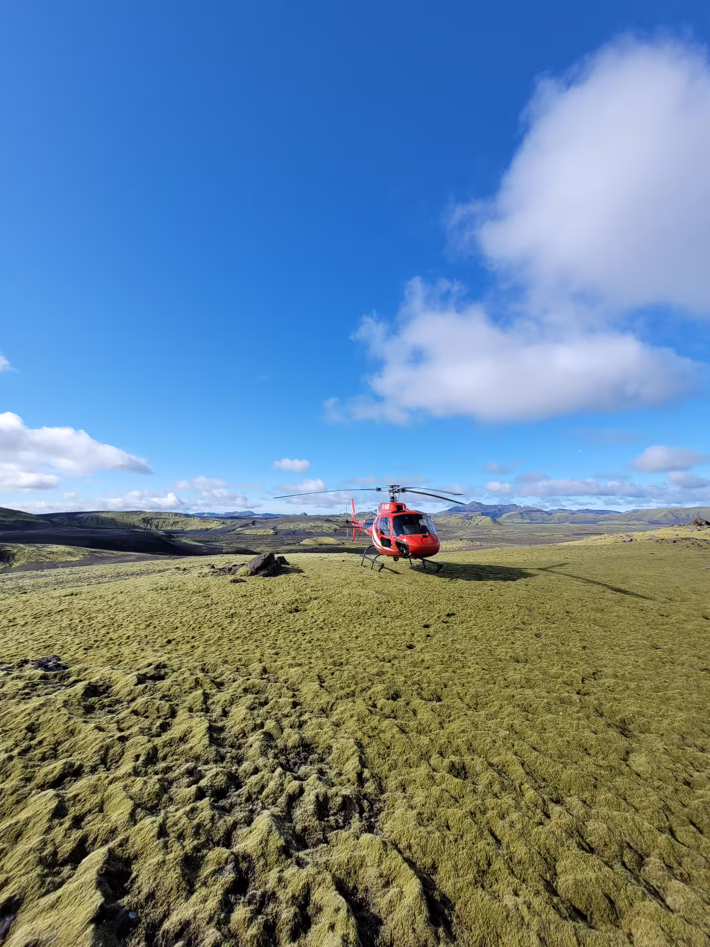 Red helicopter landed on moss-covered lava fields at Laki Craters with volcanic highlands in background, Iceland