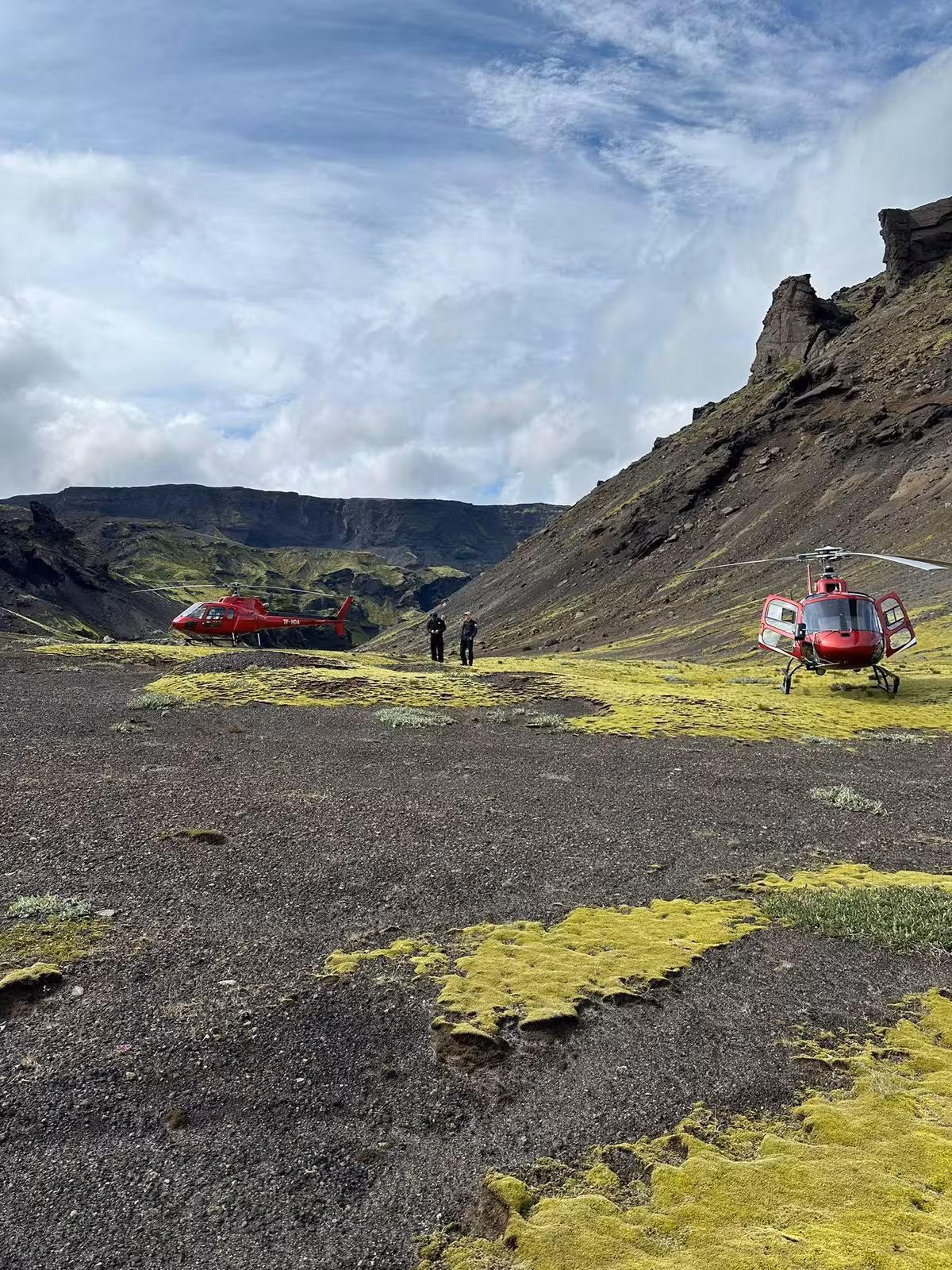 Two red helicopters landed on mossy volcanic terrain in an Icelandic highland valley with dramatic cliffs