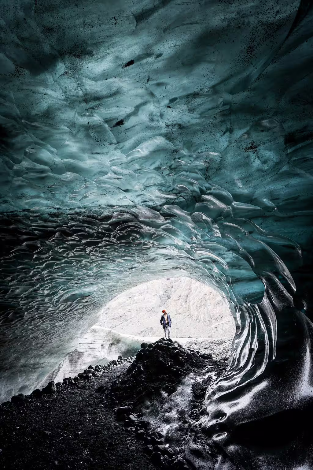 A visitor in a red helmet stands at the glowing entrance of a breathtaking blue Iceland glacier ice cave