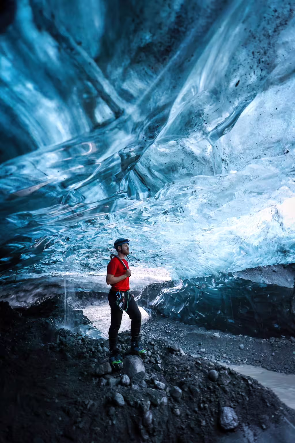 Harnessed explorer in red jacket stands beneath a dramatic swirling blue ice cave ceiling on a private glacier tour.