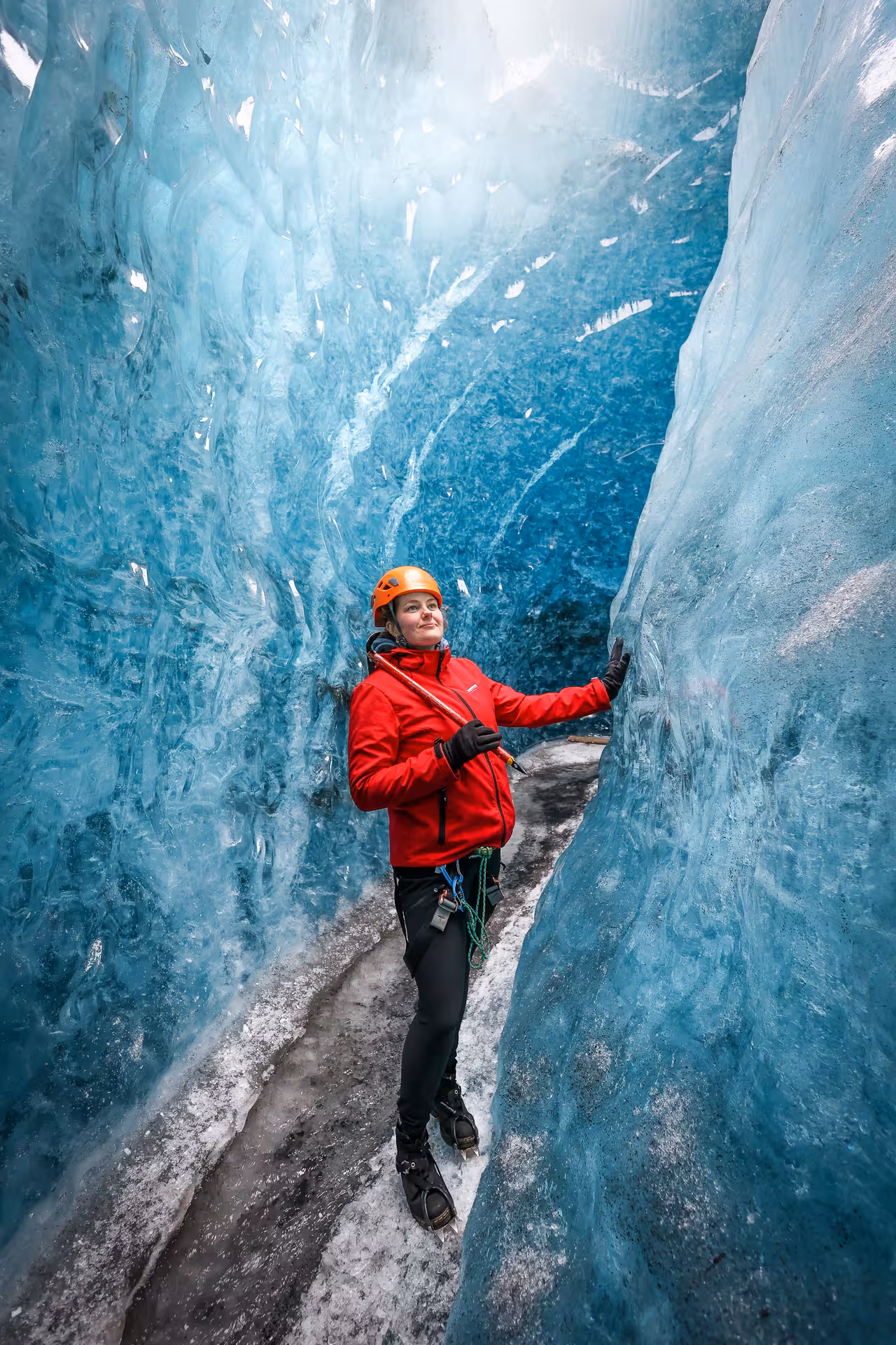 A hiker in a red jacket and orange helmet explores a stunning blue ice crevasse on Falljokull glacier guided tour Iceland