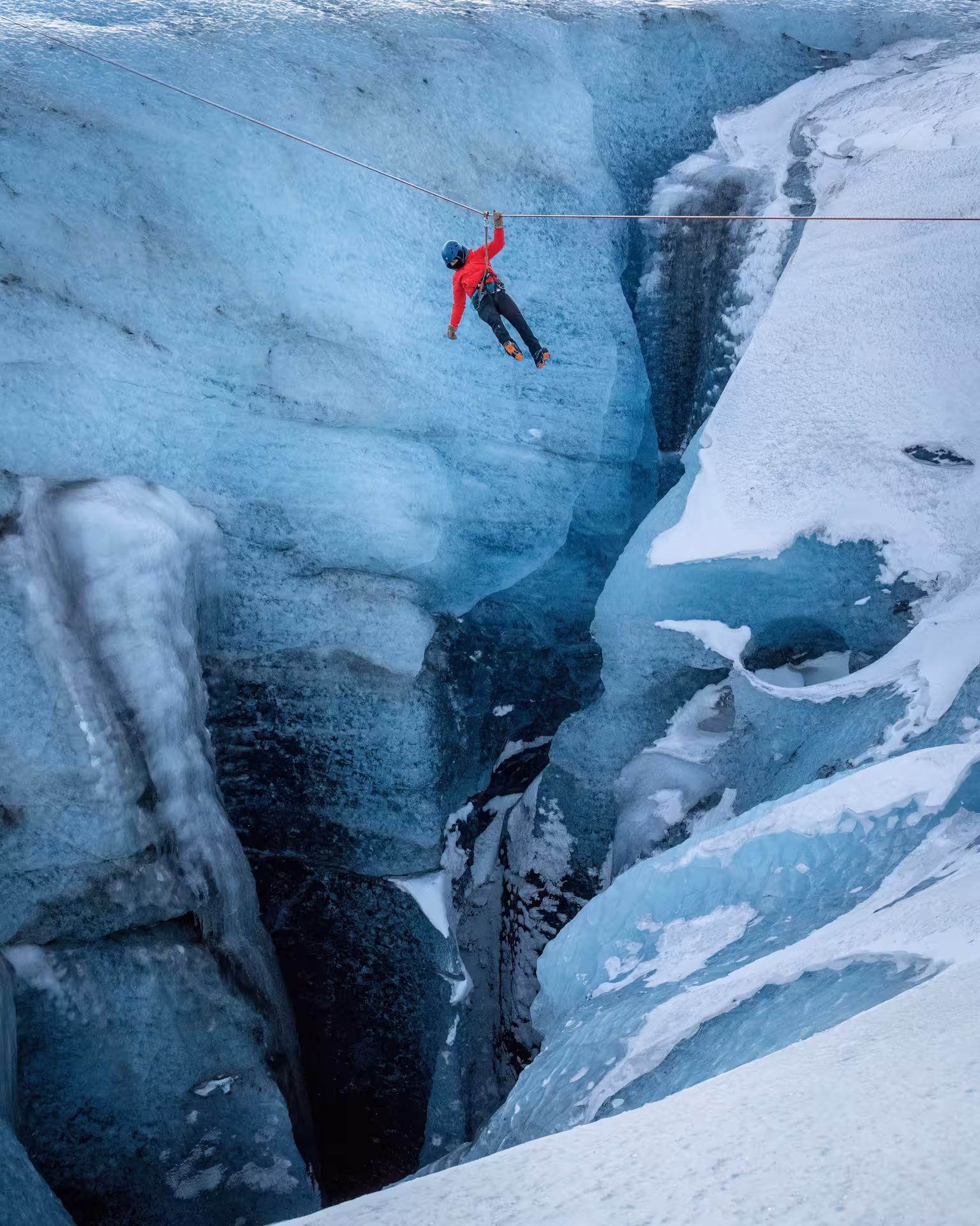 Thrill-seeker in red jacket zip lining over stunning blue glacier crevasse on Iceland winter adventure tour