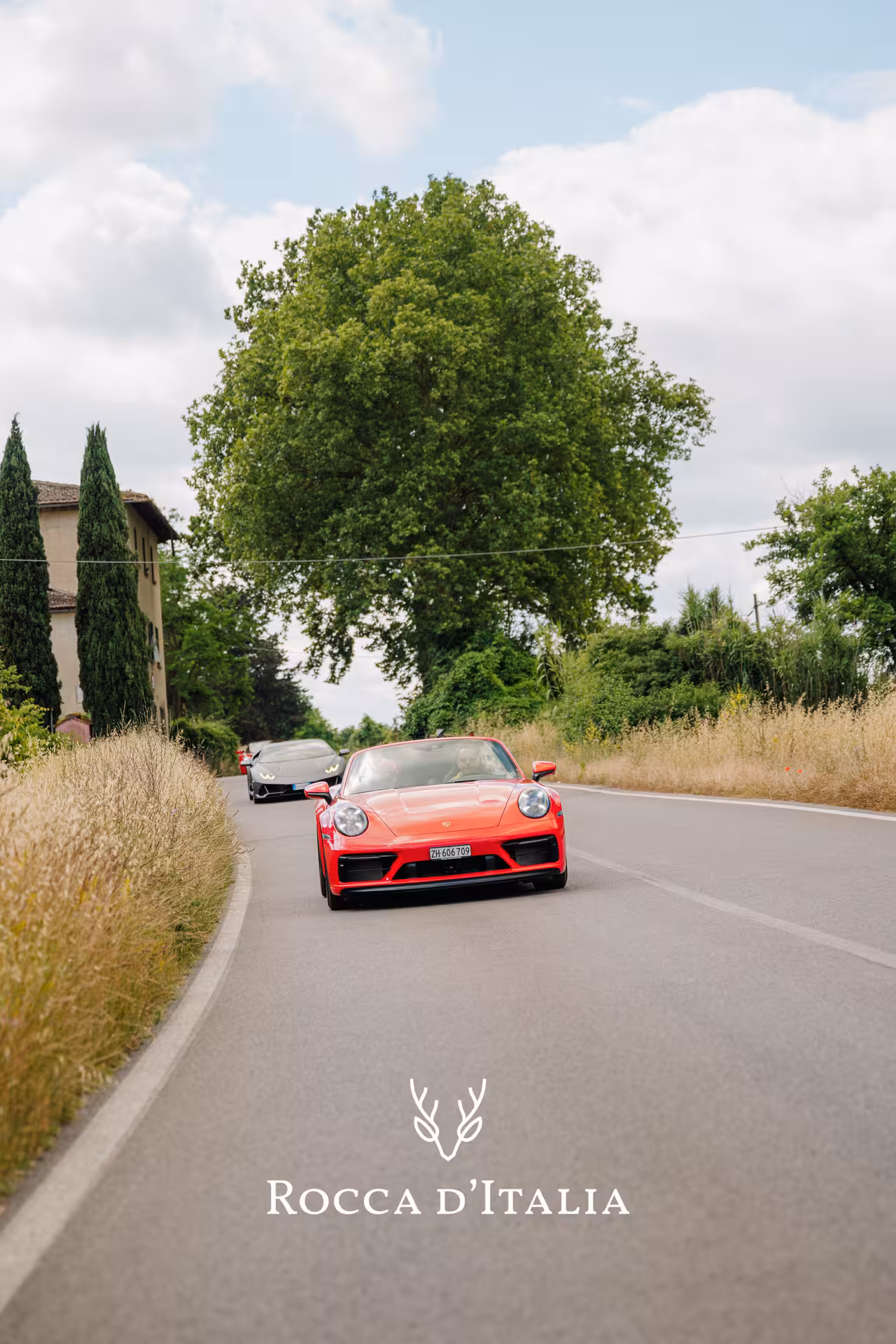 Red Porsche 911 leading a supercar convoy on a scenic Tuscan road near Siena