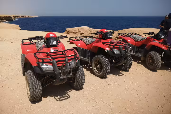 Three red quad bikes parked on rocky desert terrain overlooking the blue sea at Soma Bay