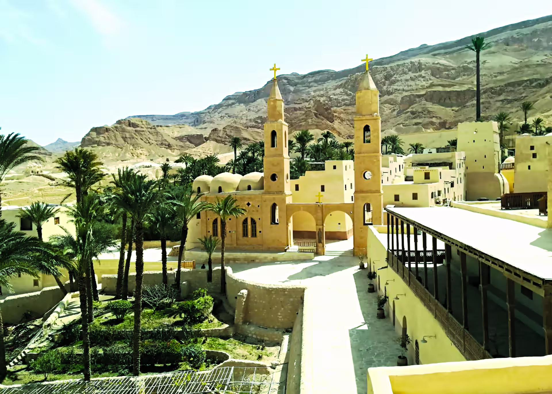 Coptic monastery with twin bell towers and golden crosses set against desert cliffs and palm trees in Egypt
