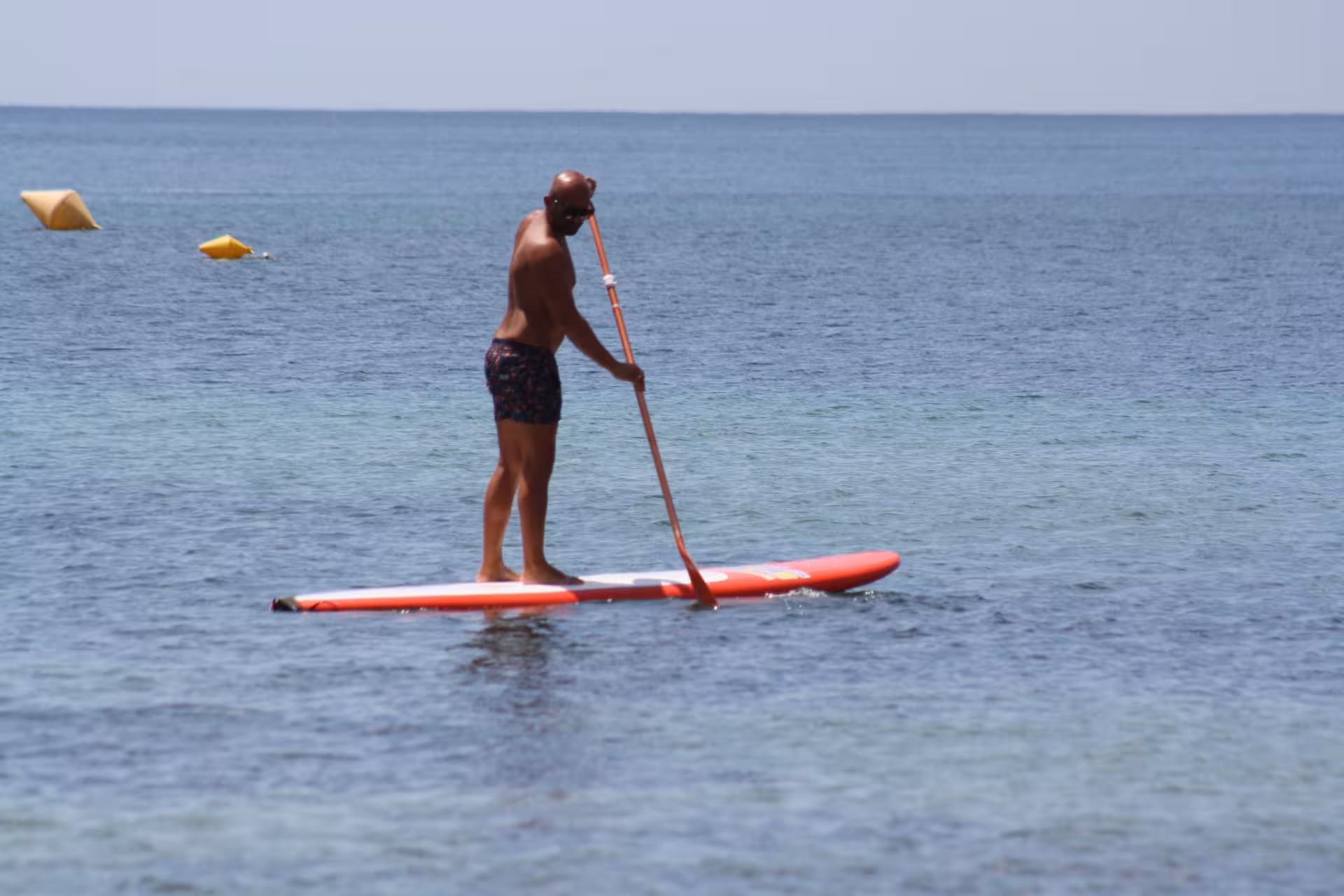 Bald man balancing on a red SUP board rental and paddling across calm coastal ocean waters