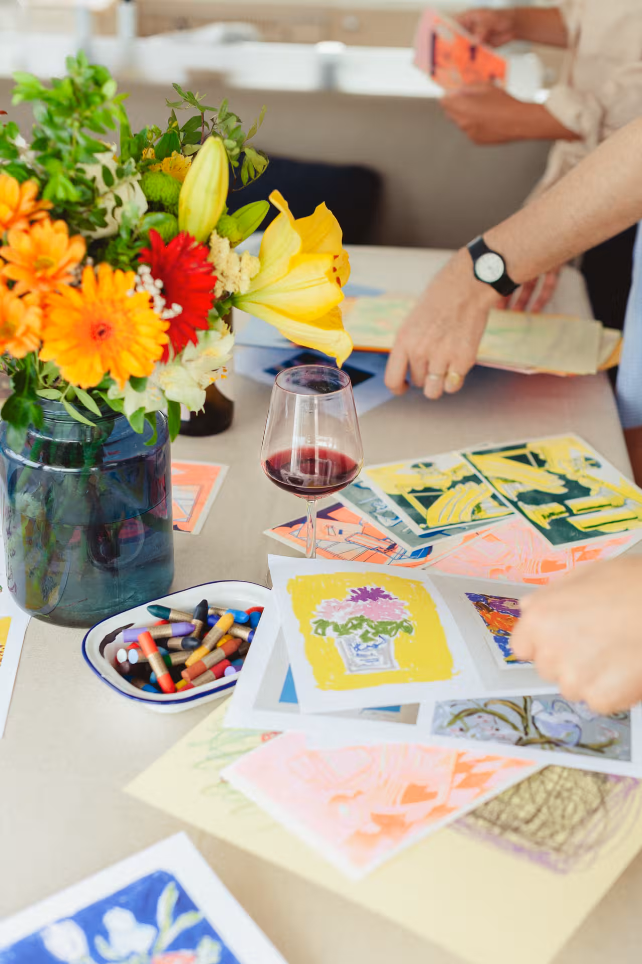 Red wine beside colorful art prints and crayons, hands creating at a Wine & Art sip and paint class