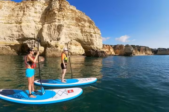 Two women on rented stand up paddleboards exploring calm turquoise waters and dramatic limestone cliffs in Algarve