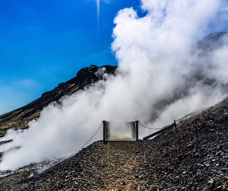 Steam rising from geothermal vents on rocky volcanic hillside with wooden bridge on Reykjadalur hike trail