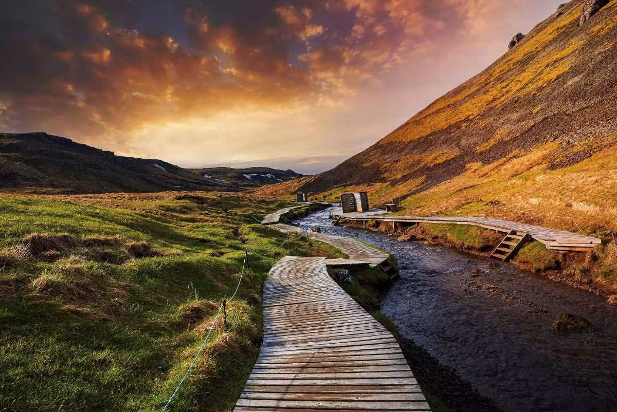 Wooden boardwalk winding along Reykjadalur hot river valley beneath dramatic sunset sky