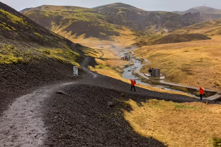 Hikers in red jackets on black volcanic trail leading to Reykjadalur geothermal hot river valley