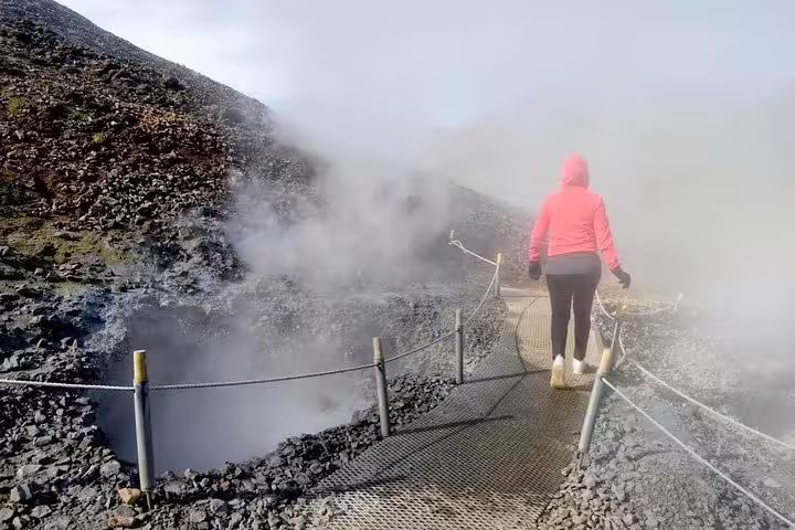 Hiker in red jacket walking boardwalk trail past steaming geothermal vents at Reykjadalur hot spring