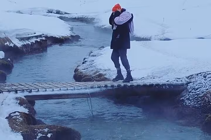 Person standing on wooden bridge over geothermal stream at Reykjadalur Hot Springs in winter snow