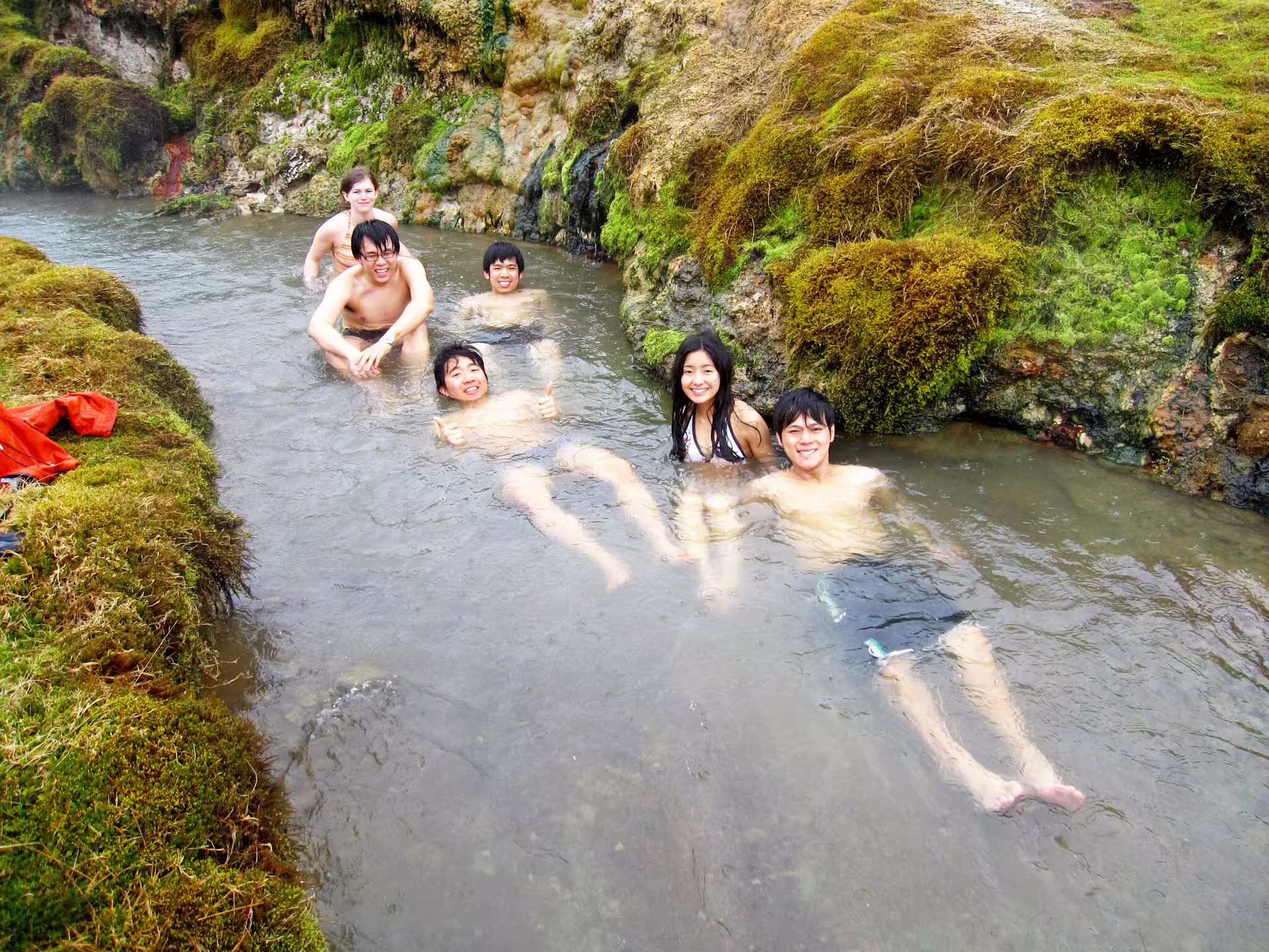 Group enjoying a relaxing soak in Reykjadalur Valley's geothermal hot river, surrounded by lush green moss and natural beauty.