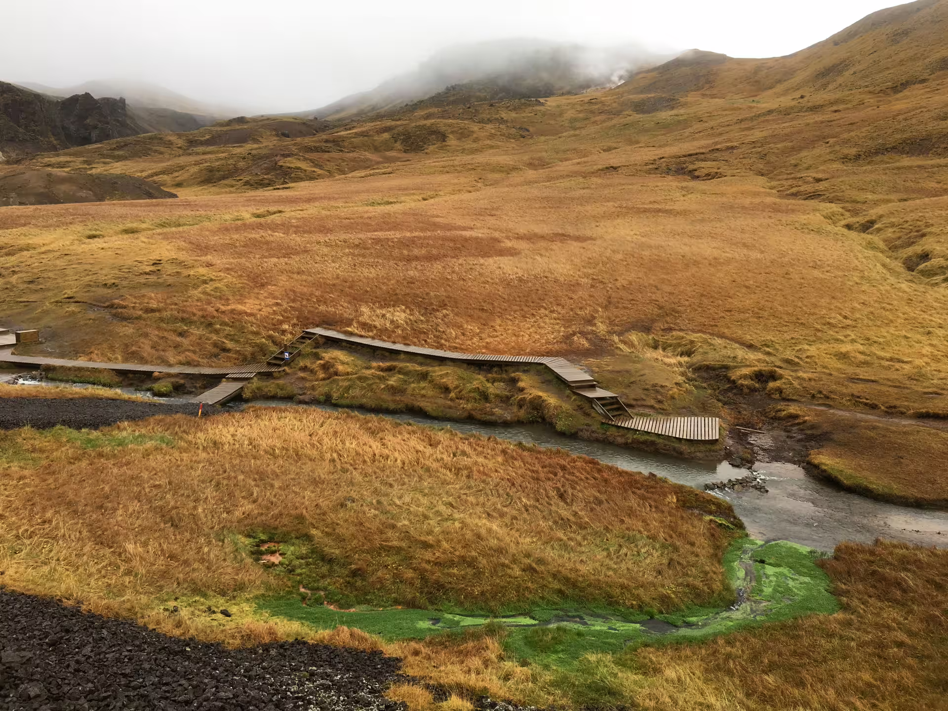 Scenic view of Reykjadalur Valley's geothermal landscape with a wooden path over the hot river amidst autumn foliage.