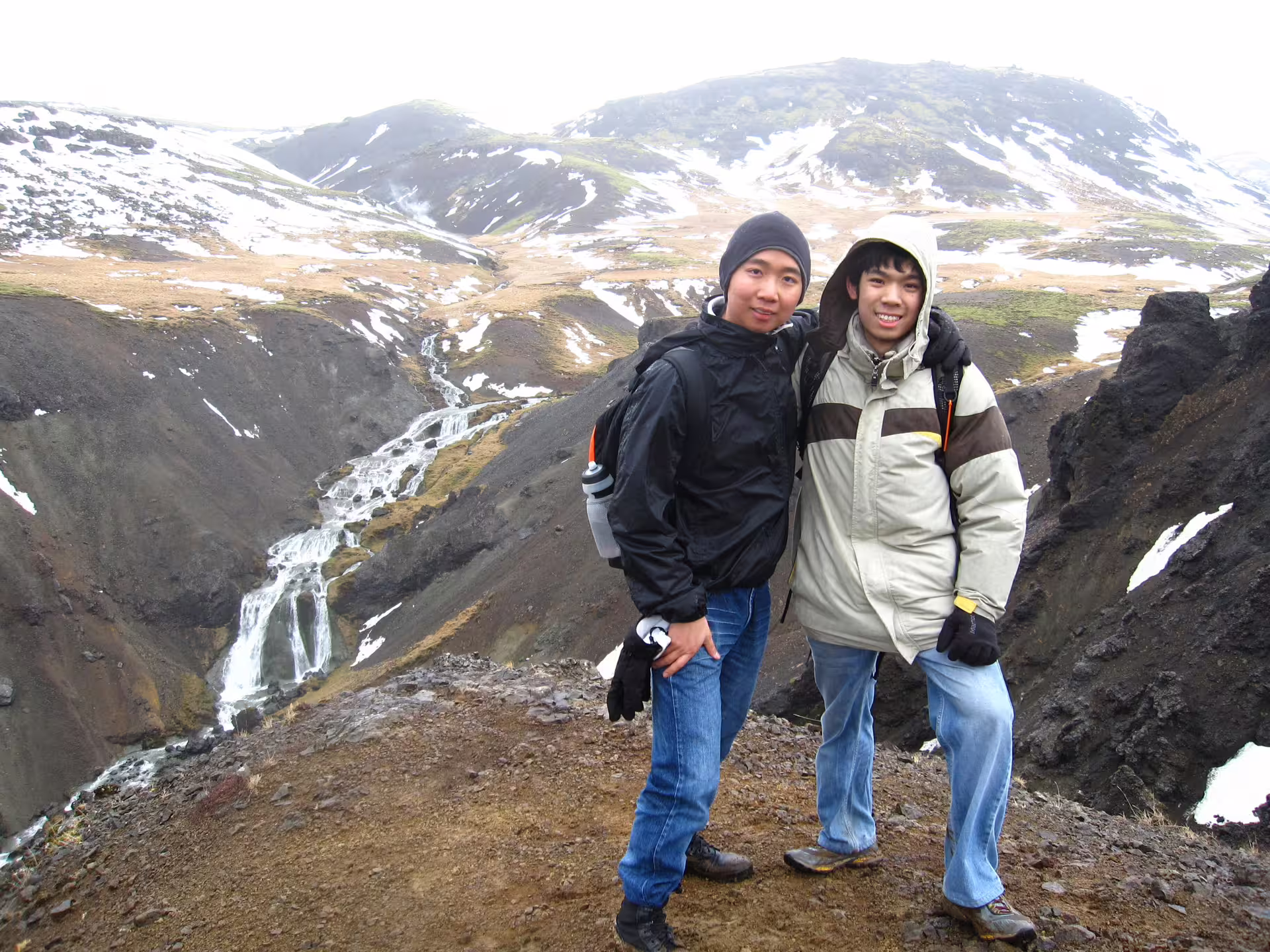 Two hikers in winter gear enjoy a scenic view of Reykjadalur Valley's snowy landscape and cascading waterfall in Iceland.