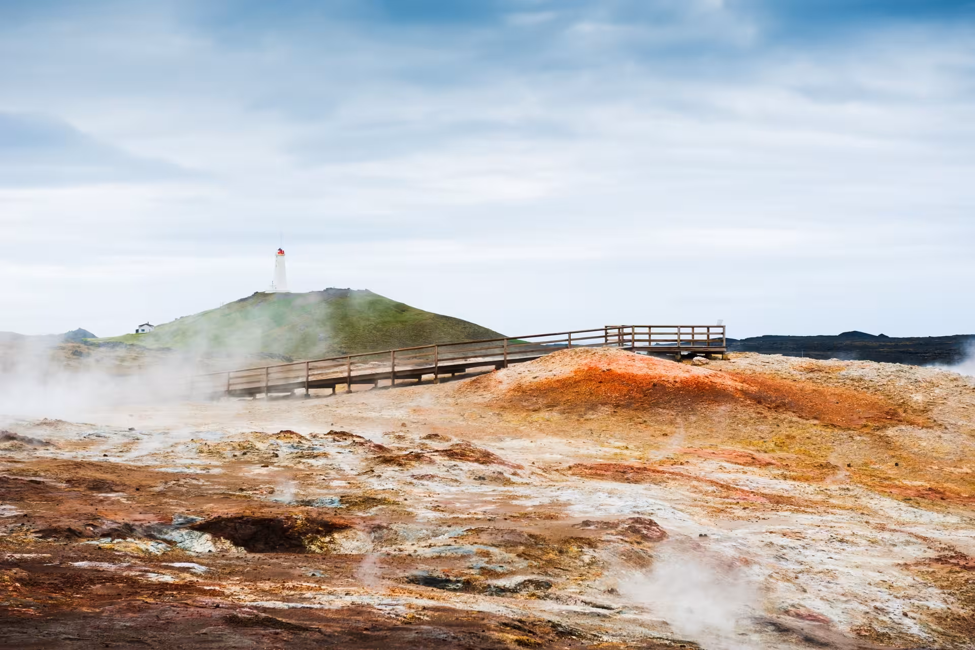 Steaming geothermal landscape with colorful mineral deposits, wooden boardwalk, and lighthouse at Reykjanes Geopark, Iceland