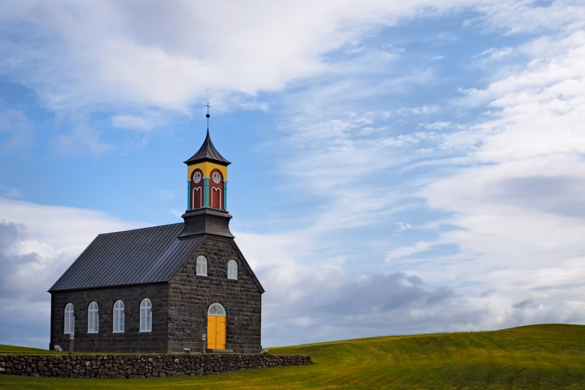 Historic dark stone church with colorful bell tower on green hillside in Reykjanes Iceland