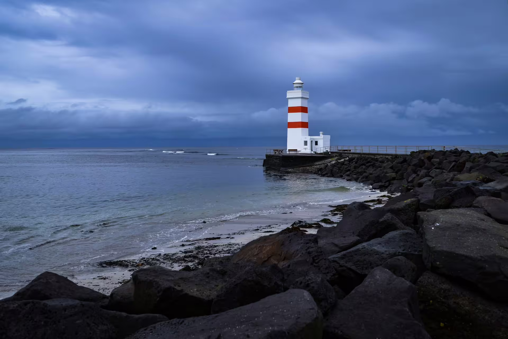 White and red striped lighthouse on rocky Reykjanes Peninsula coastline under dramatic cloudy sky