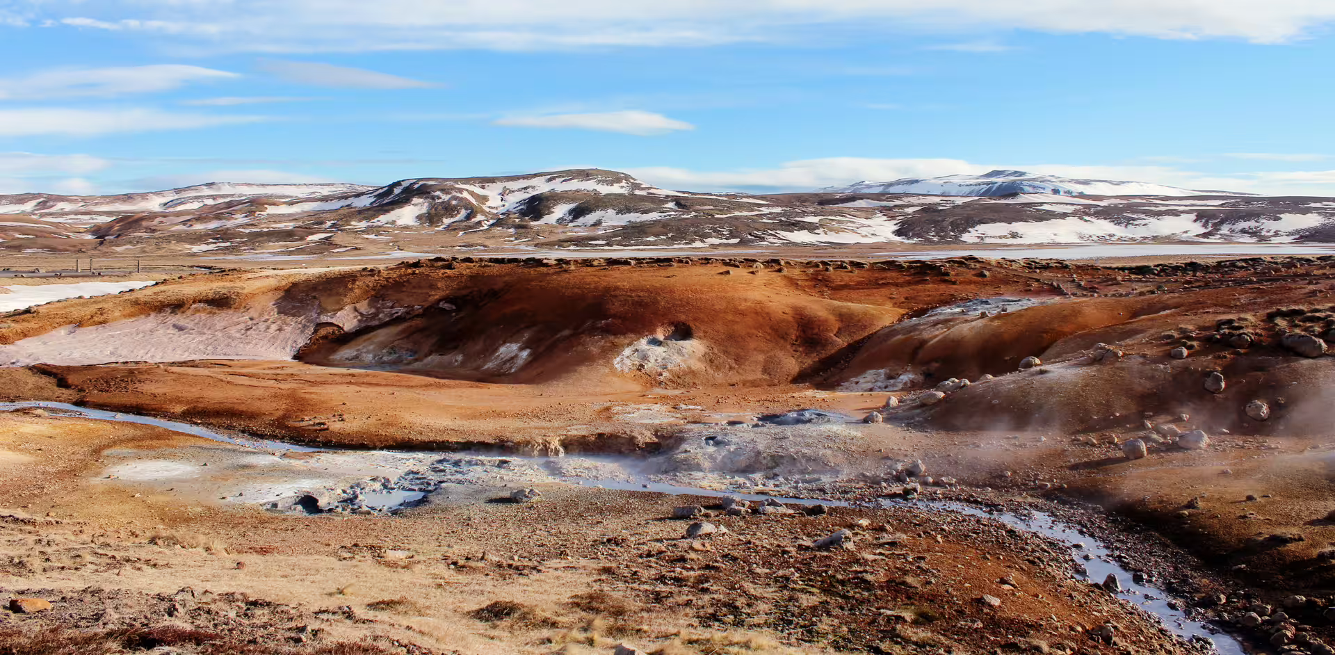 Steaming geothermal vents and rust-colored volcanic terrain near Reykjanes with snow-capped hills in background