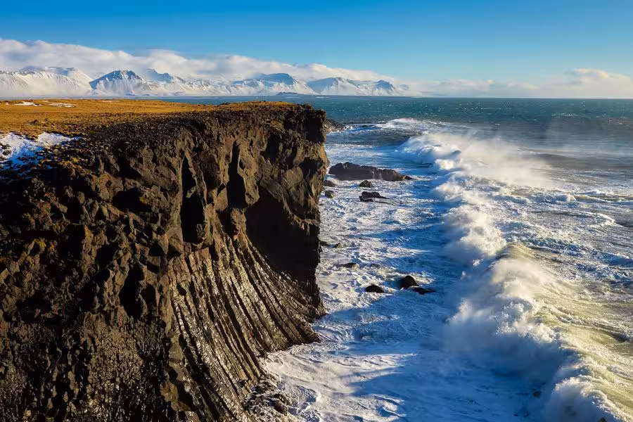 Rocky basalt sea cliffs with crashing waves and snow-capped mountains in Reykjanes Iceland