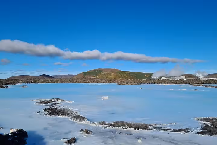 Milky blue geothermal lagoon with rocky shores, steam rising near buildings, and green volcanic hills under blue sky in Iceland