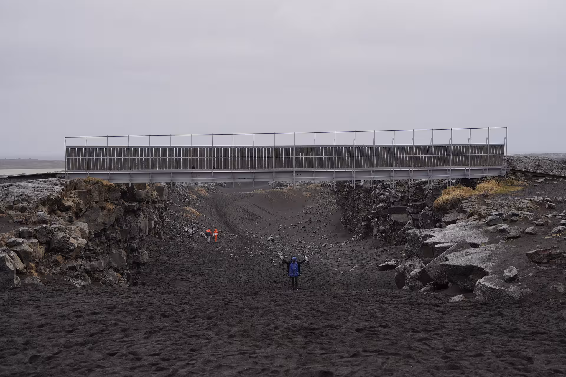Person standing in volcanic black sand canyon beneath metal bridge on Reykjanes Peninsula, Iceland