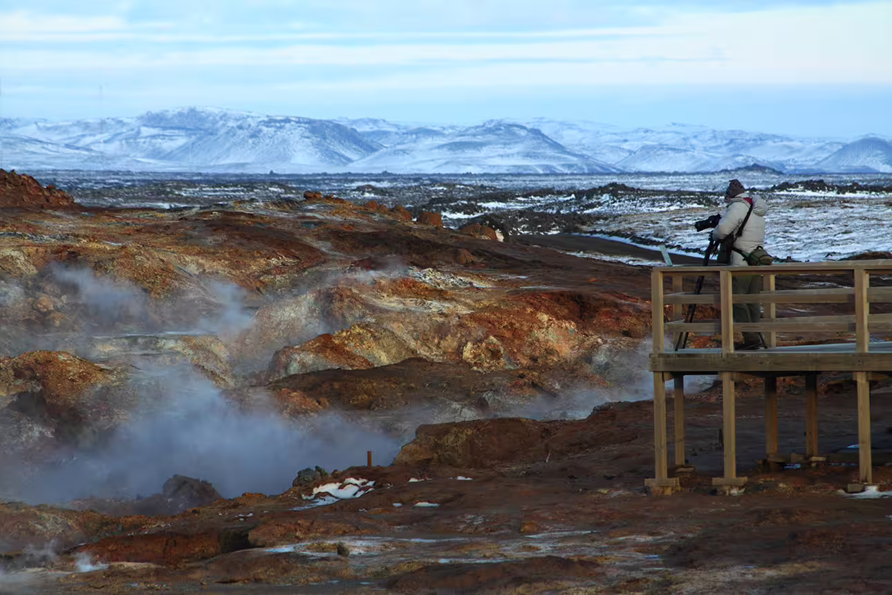 Photographer on wooden platform overlooking steaming geothermal hot springs and red volcanic rocks on Reykjanes Peninsula