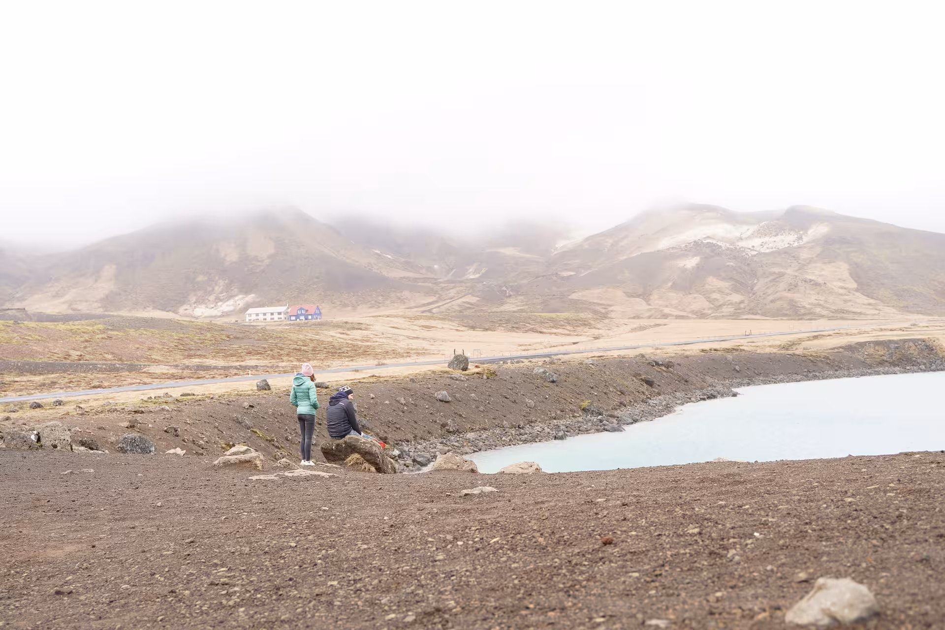 Two travelers sitting by a milky blue geothermal lake on the Reykjanes Peninsula with misty mountains behind