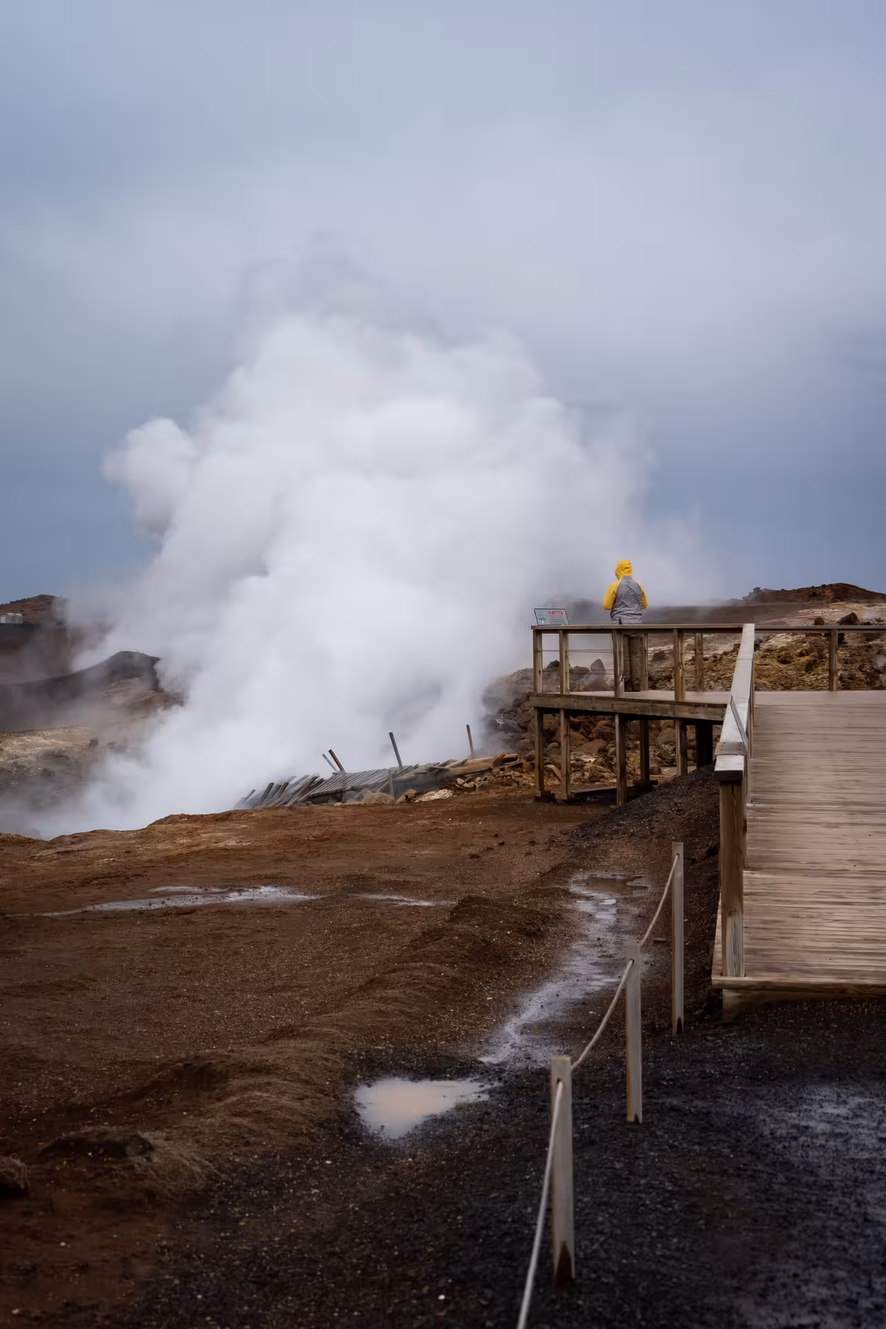 Visitor in yellow jacket on wooden viewing platform overlooking steaming geothermal vents on Reykjanes Peninsula