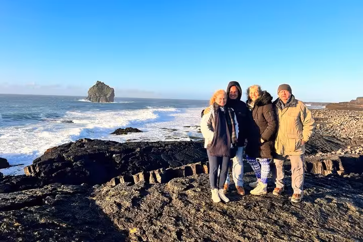 Four tourists posing on rocky Reykjanes Peninsula coastline with crashing waves and sea stack in background
