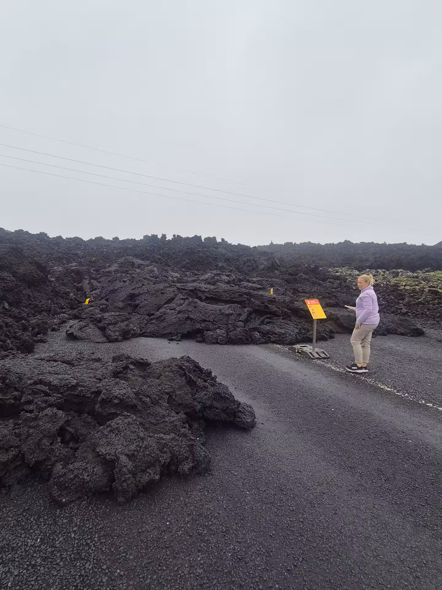 Woman reading warning sign beside hardened lava flow covering road on Reykjanes Peninsula, Iceland