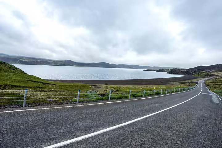 Winding road along Reykjanes Peninsula with lava fields and calm lake under cloudy skies