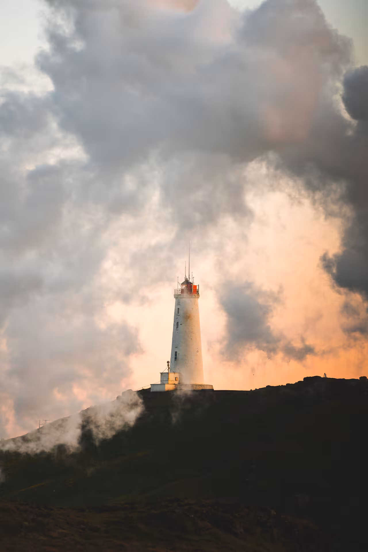 White lighthouse on Reykjanes Peninsula hillside with dramatic sunset clouds and geothermal steam rising