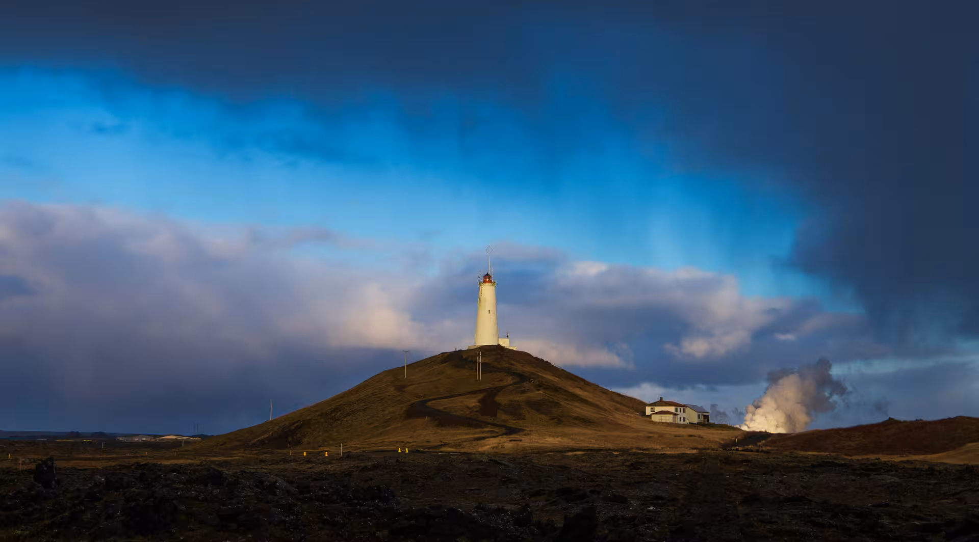 White lighthouse atop a grassy hill on Reykjanes Peninsula with dramatic blue sky and geothermal steam