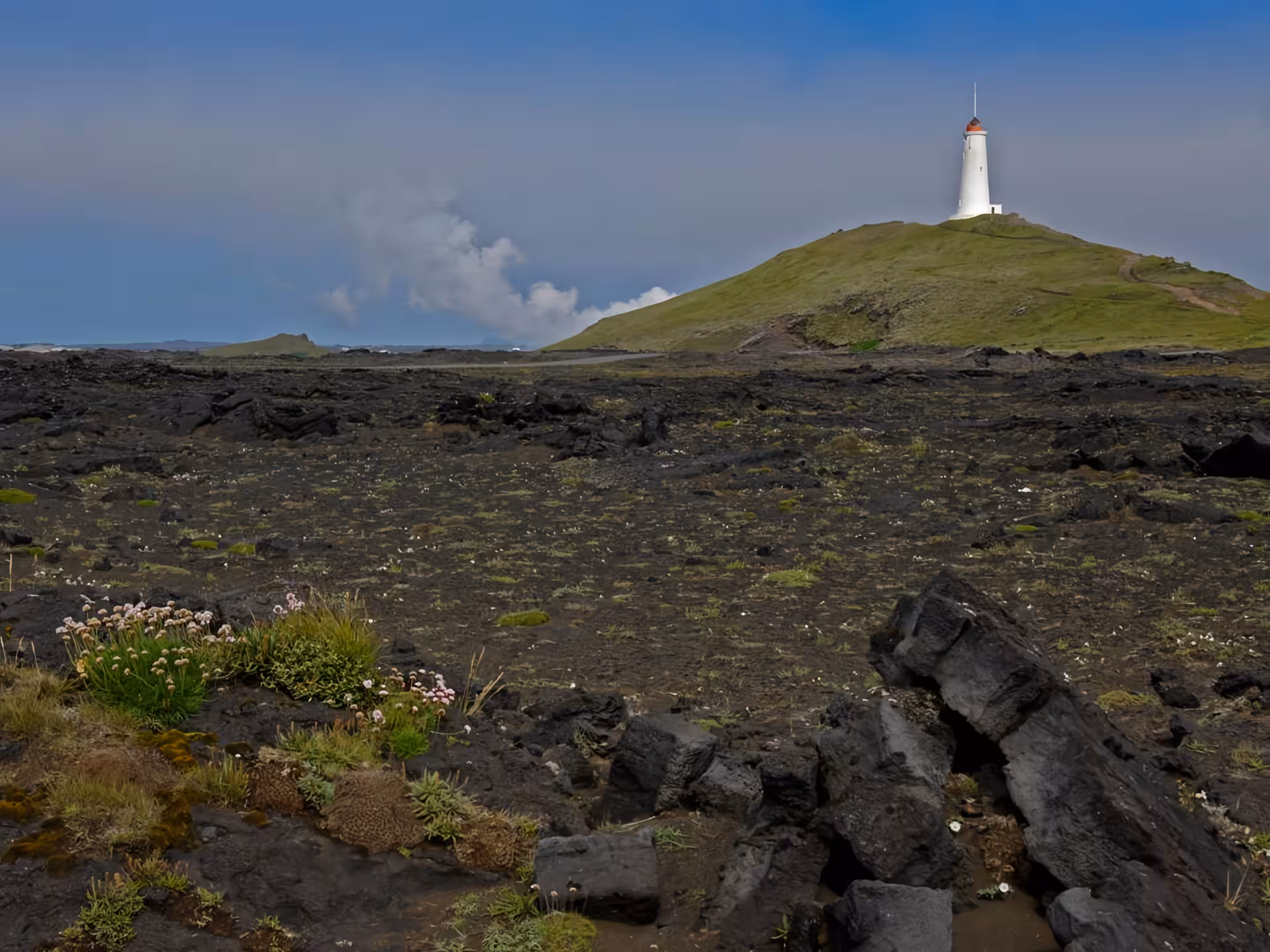 White lighthouse on green hill overlooking black lava fields on Reykjanes Peninsula, Iceland