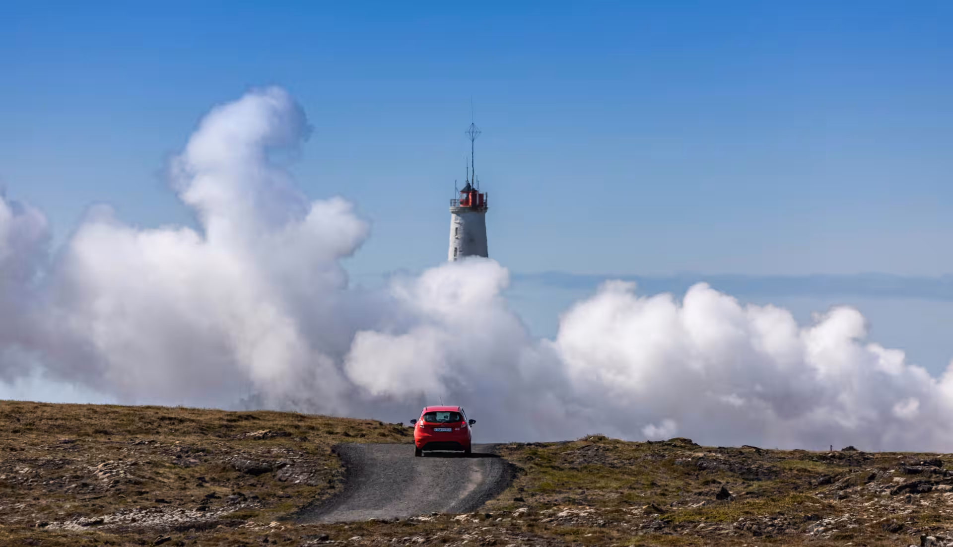 Red car driving toward a lighthouse emerging from clouds on the Reykjanes Peninsula, Iceland