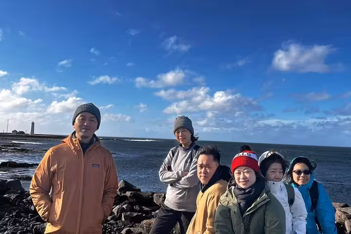 Group of six tourists posing on rocky Reykjanes Peninsula coastline with lighthouse and blue sky