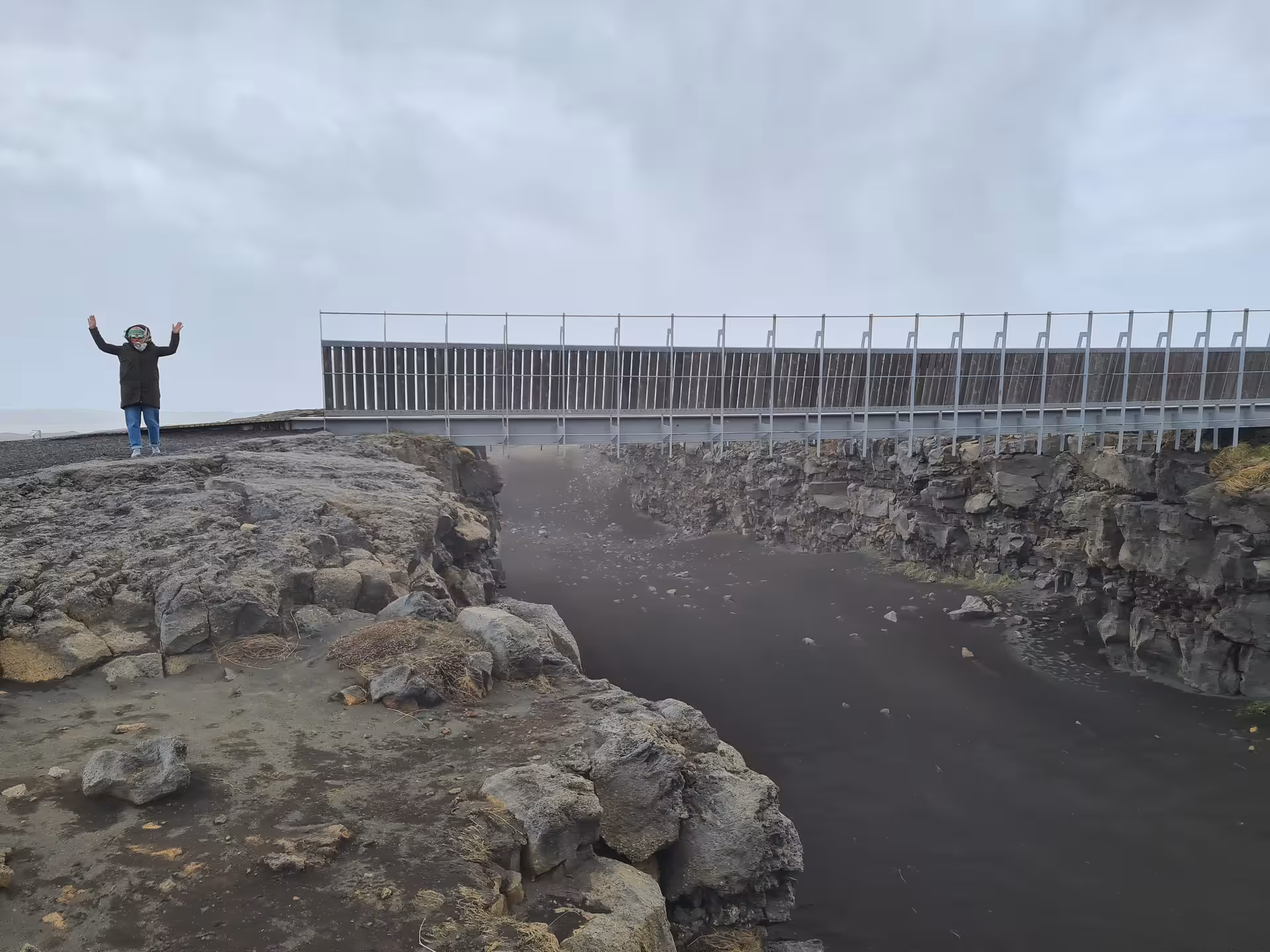 Tourist with raised arms standing at tectonic plate fissure on Reykjanes Peninsula, Iceland, with bridge overhead