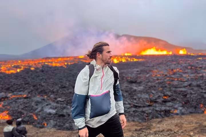 Hiker standing on volcanic lava field with erupting volcano glowing orange in background, Reykjanes Peninsula