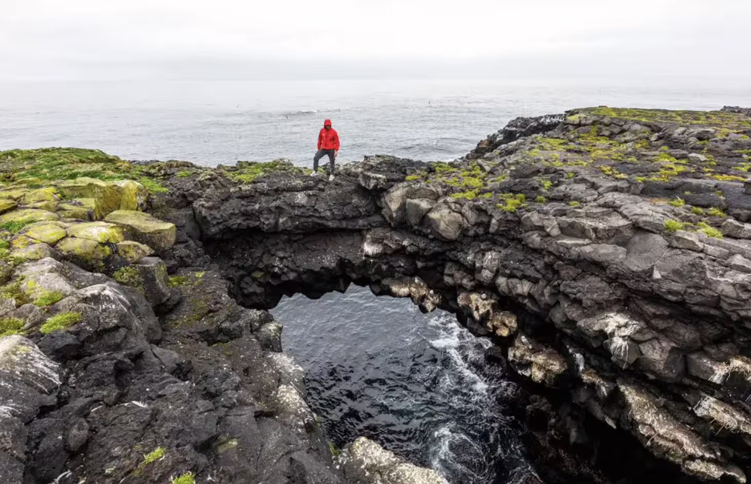 Person in red jacket standing on volcanic rock arch over ocean pool on Reykjanes Peninsula Iceland