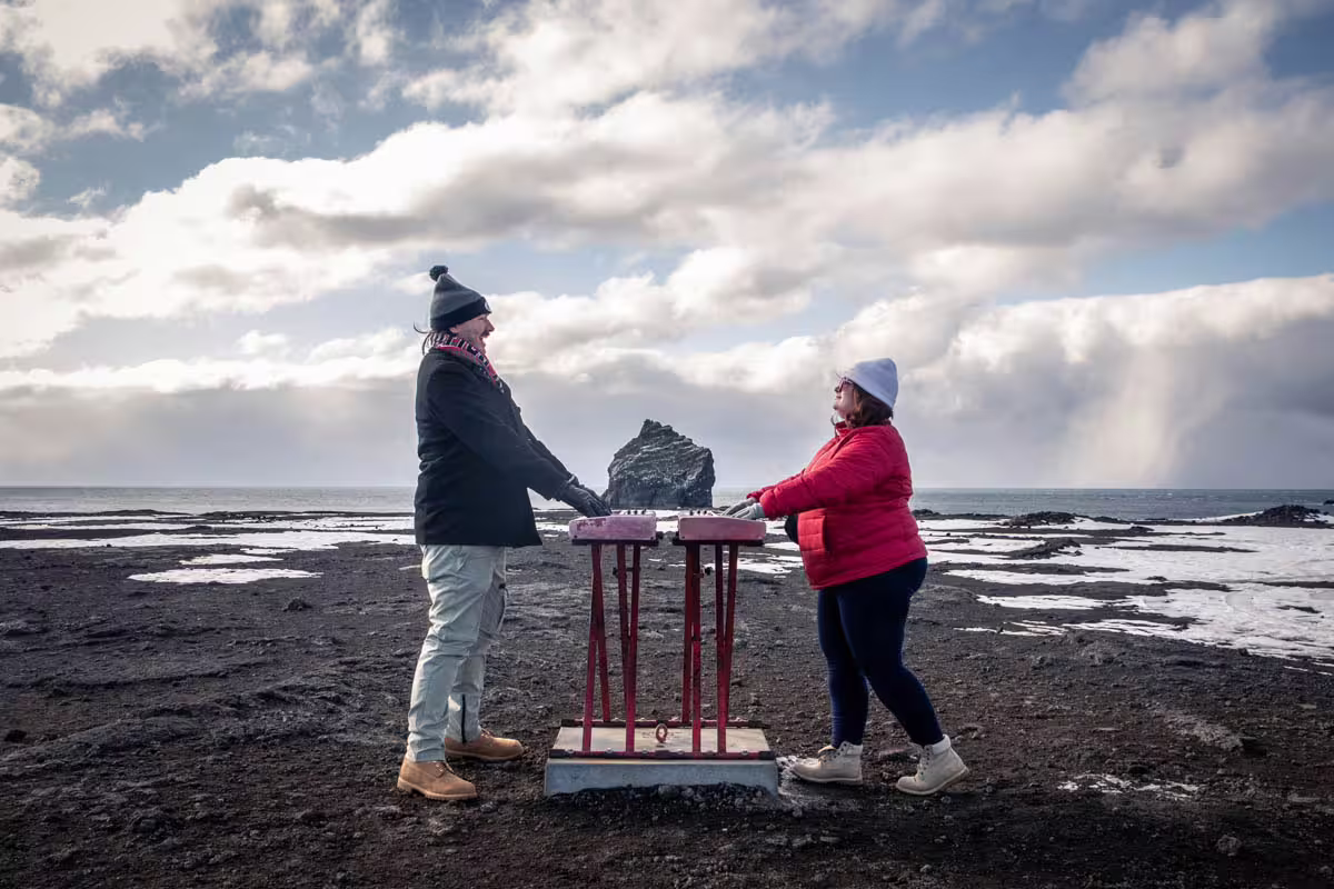 Two hikers in winter gear at a red gate on Iceland's black volcanic Reykjanes Peninsula coastline