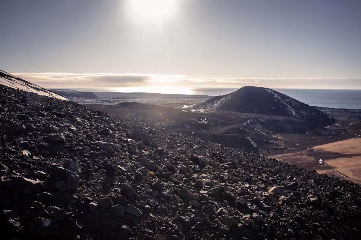 Rocky volcanic landscape on Reykjanes Peninsula with dark lava fields, volcanic cone, and ocean in background