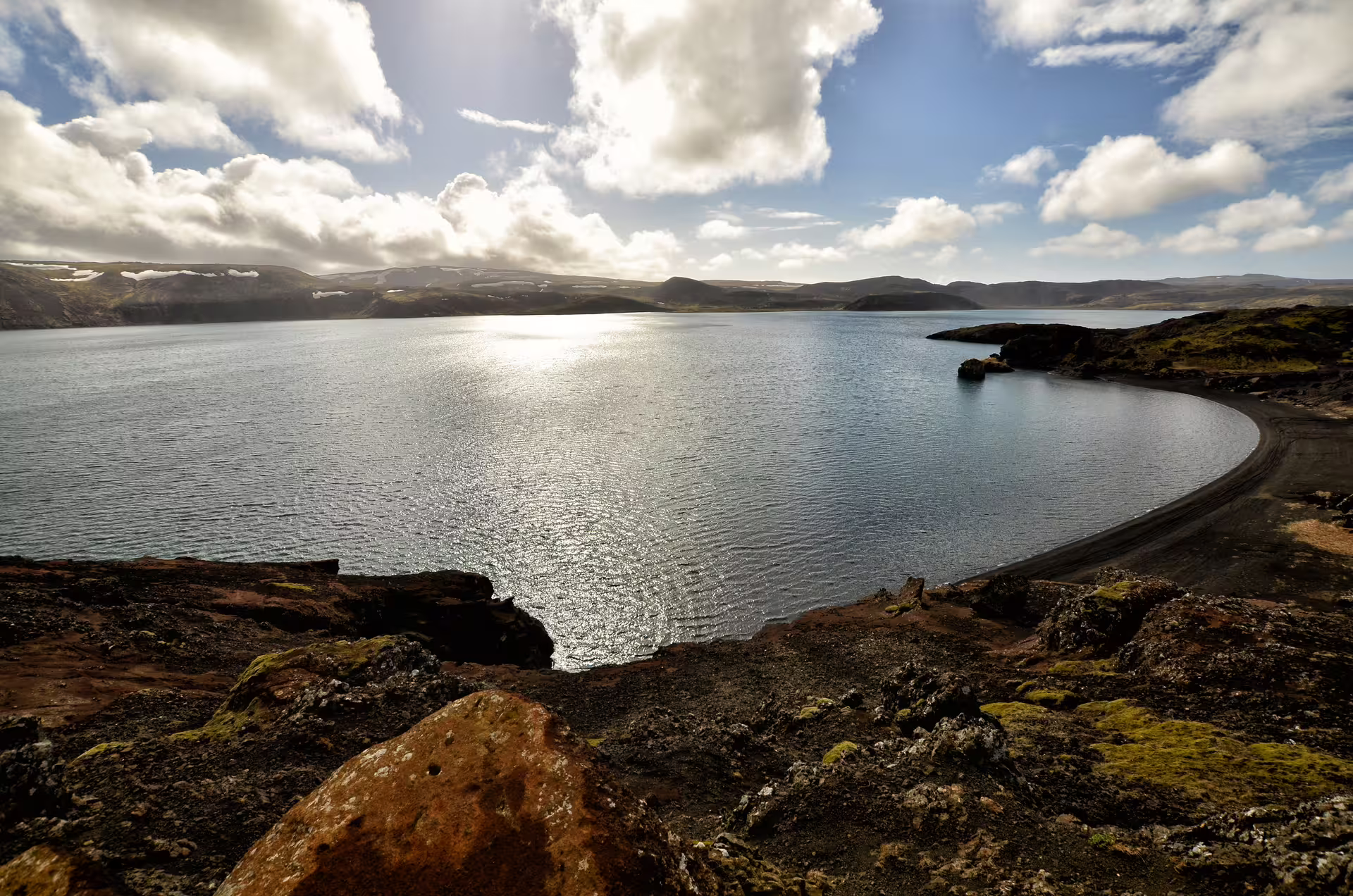 Volcanic lake with black sand shore, lava rocks, and green moss under dramatic cloudy sky in Reykjanes, Iceland