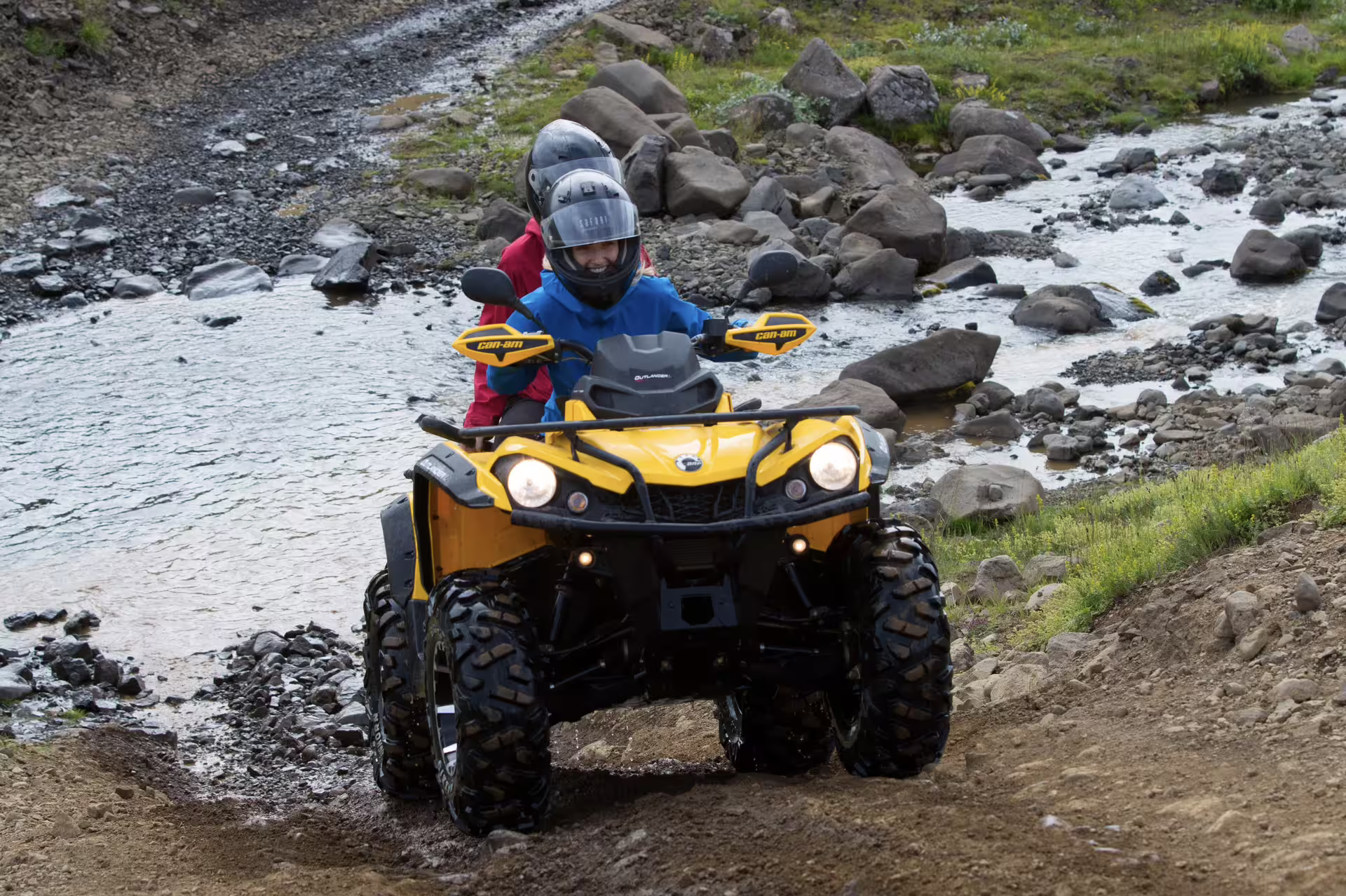Two riders in helmets on a yellow Can-Am ATV navigating rocky river terrain near Reykjavik