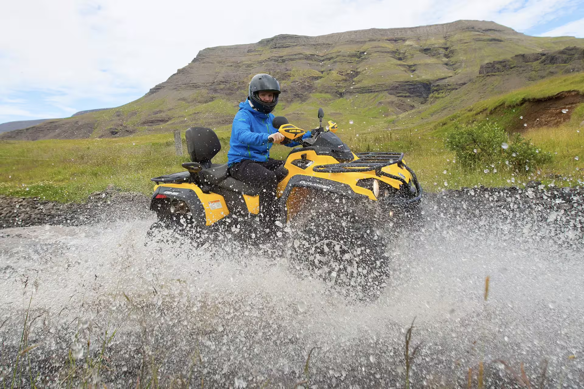 Rider on yellow Can-Am Outlander ATV splashing through river crossing with green Icelandic mountains behind