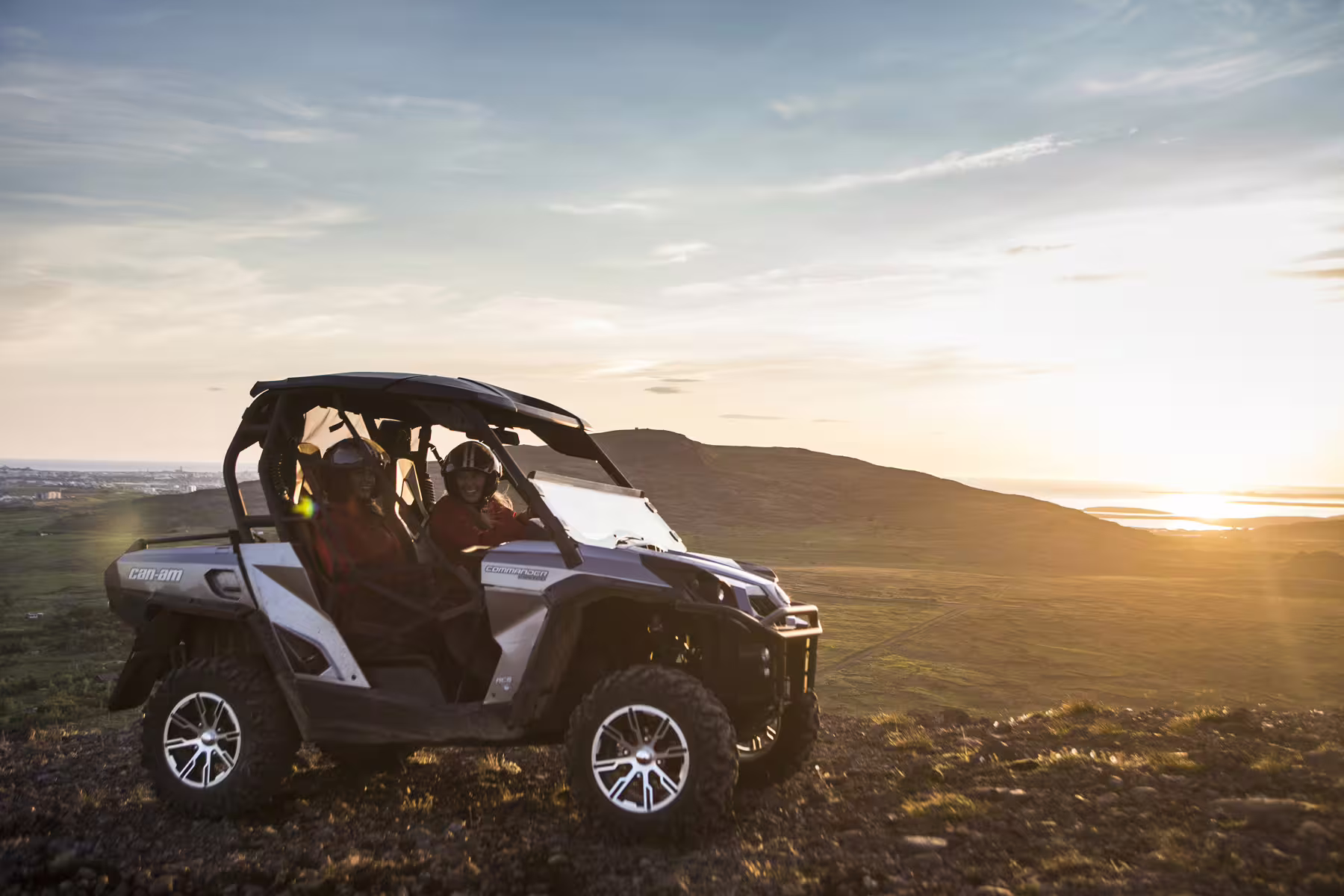 Two riders in helmets sitting in a white Can-Am Commander buggy on a hilltop at sunset in Reykjavik