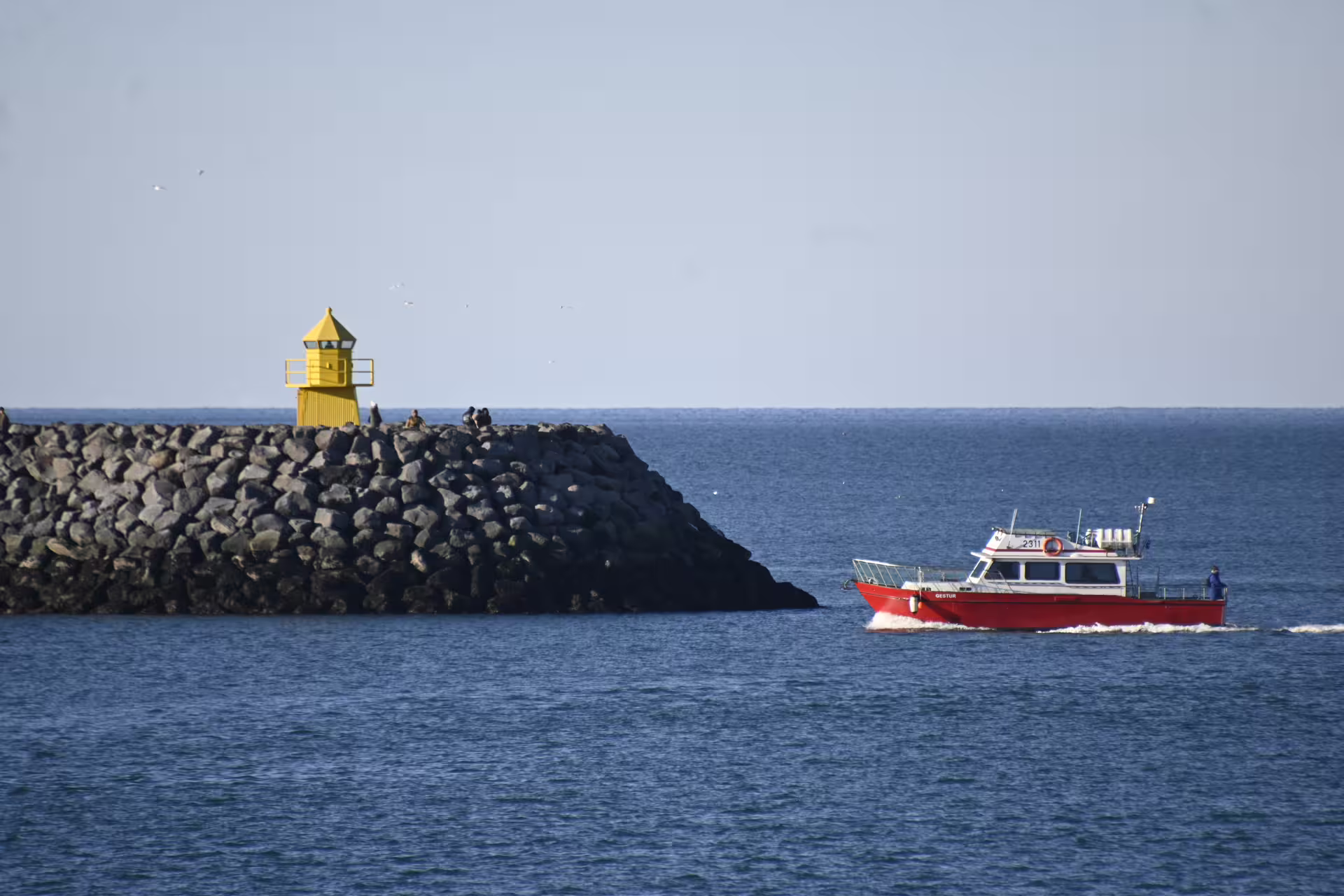 Red ferry boat passing yellow lighthouse on rocky breakwater in Reykjavik harbor