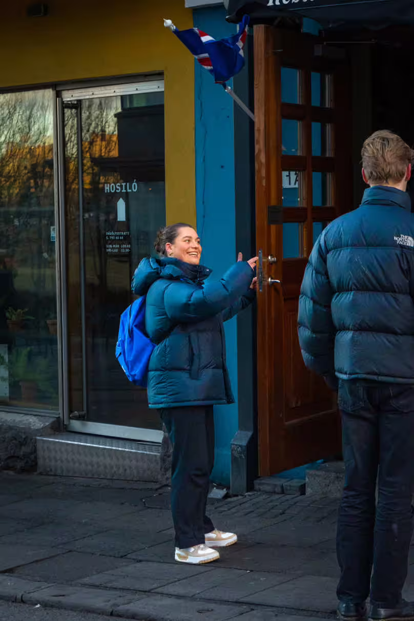 Smiling woman in blue puffer jacket pointing at Icelandic flag outside Reykjavik restaurant