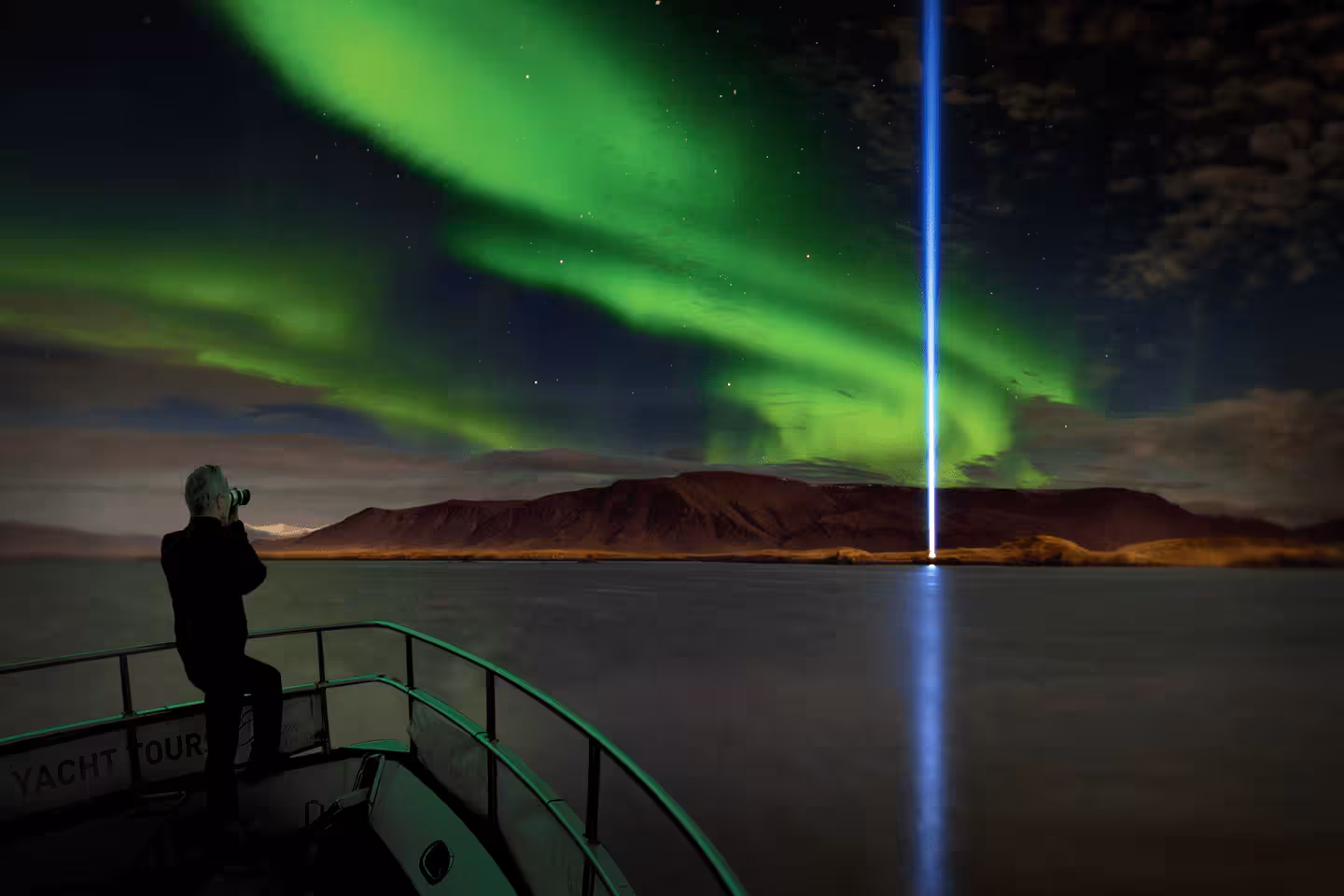 Photographer on yacht tour bow capturing northern lights and blue light beam over Reykjavik harbor at night